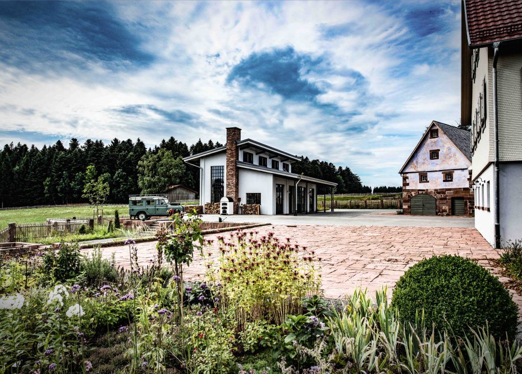 Der Hof der Destillerie in Loßburg: ein weißes Gebäude mit hohem Kamin und Werkstattfenstern, davor ein gepflasterter Platz mit Kräuterbeet, dahinter Wald