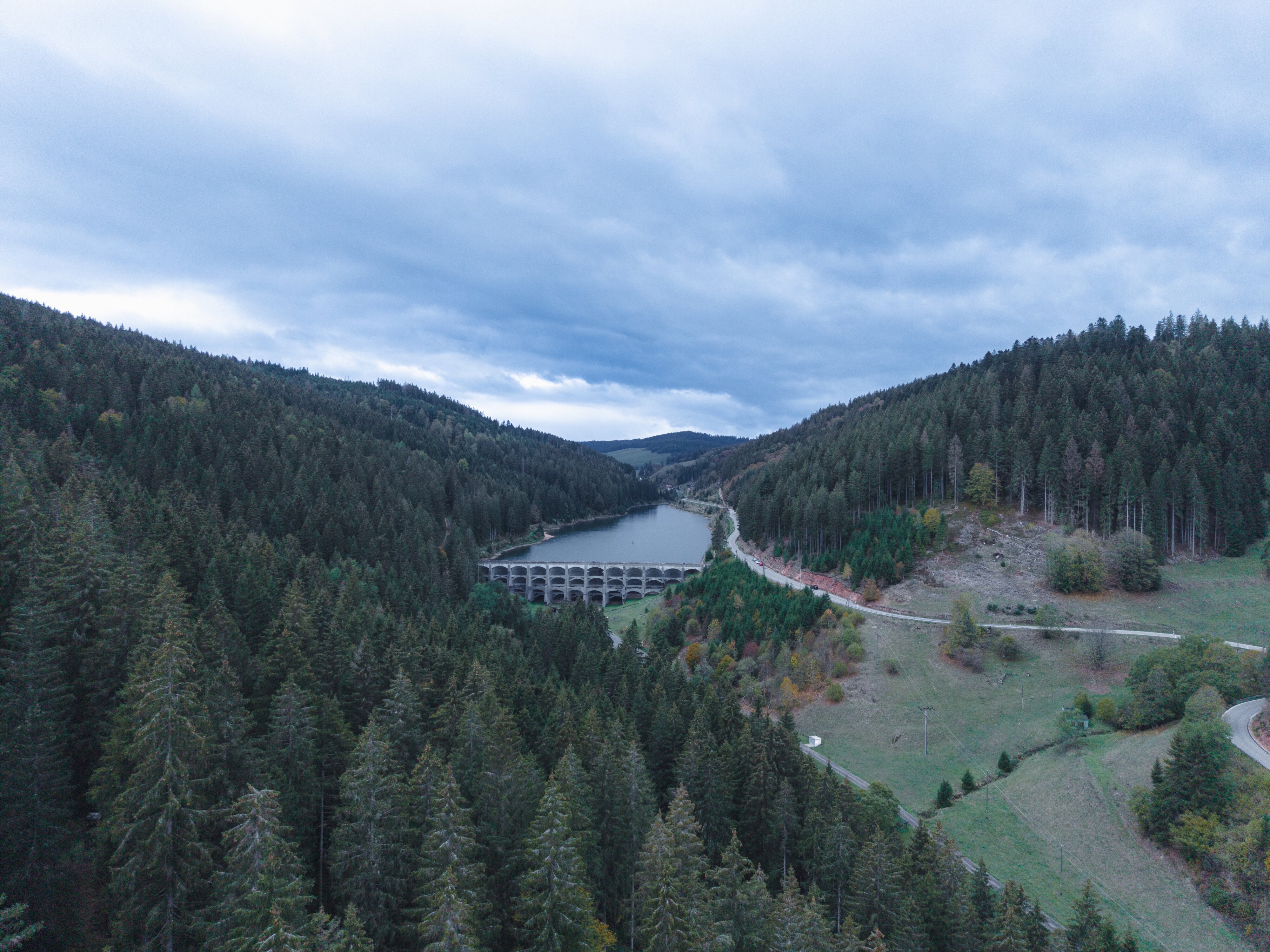 Das bewaldete Linachtal bei Vöhrenbach aus großer Höhe, in der Mitte klein die Staumauer mit See unter grauem Abendhimmel.