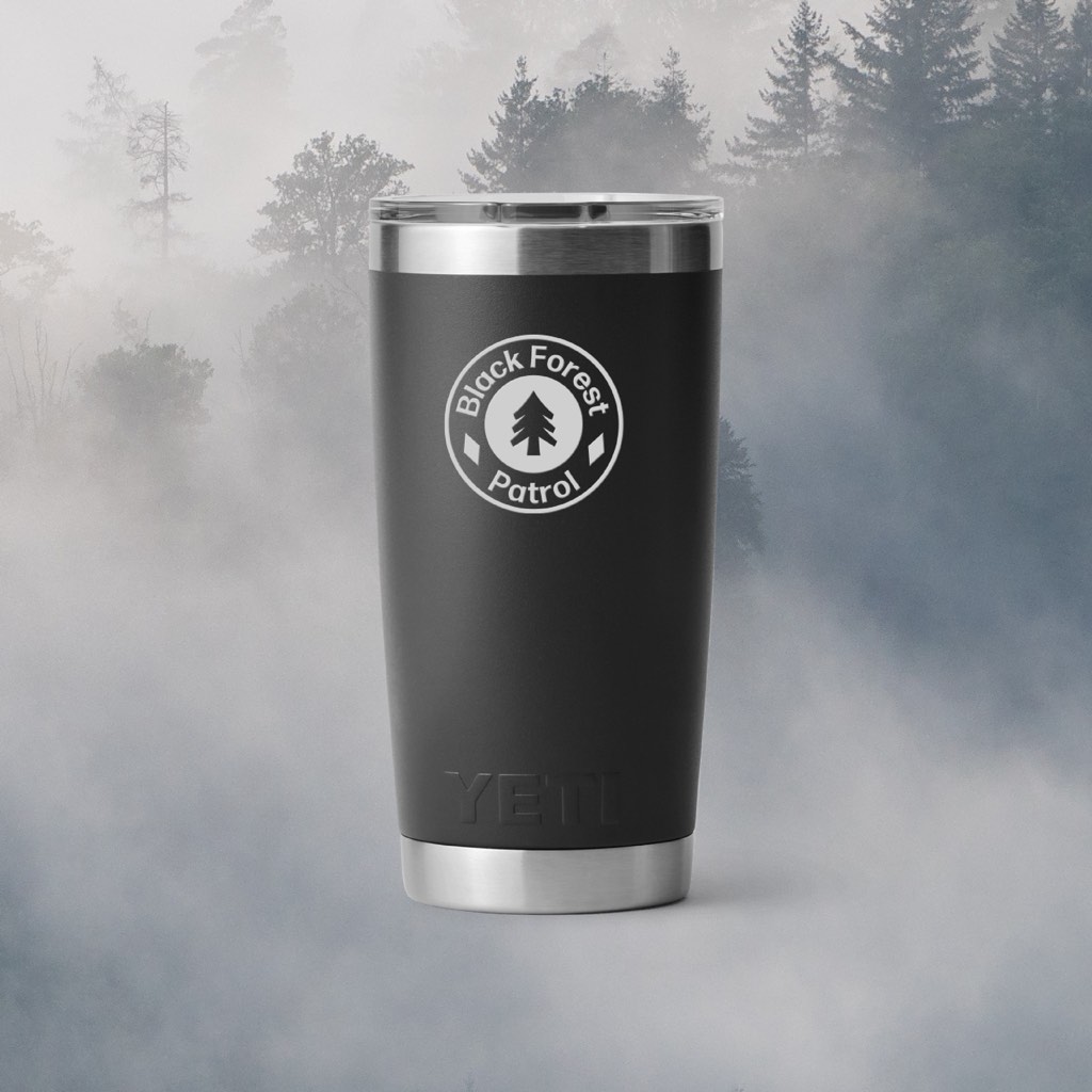 Schwarzer Edelstahlbecher mit Deckel und rundem weißem Zeichen, vor nebligem Wald