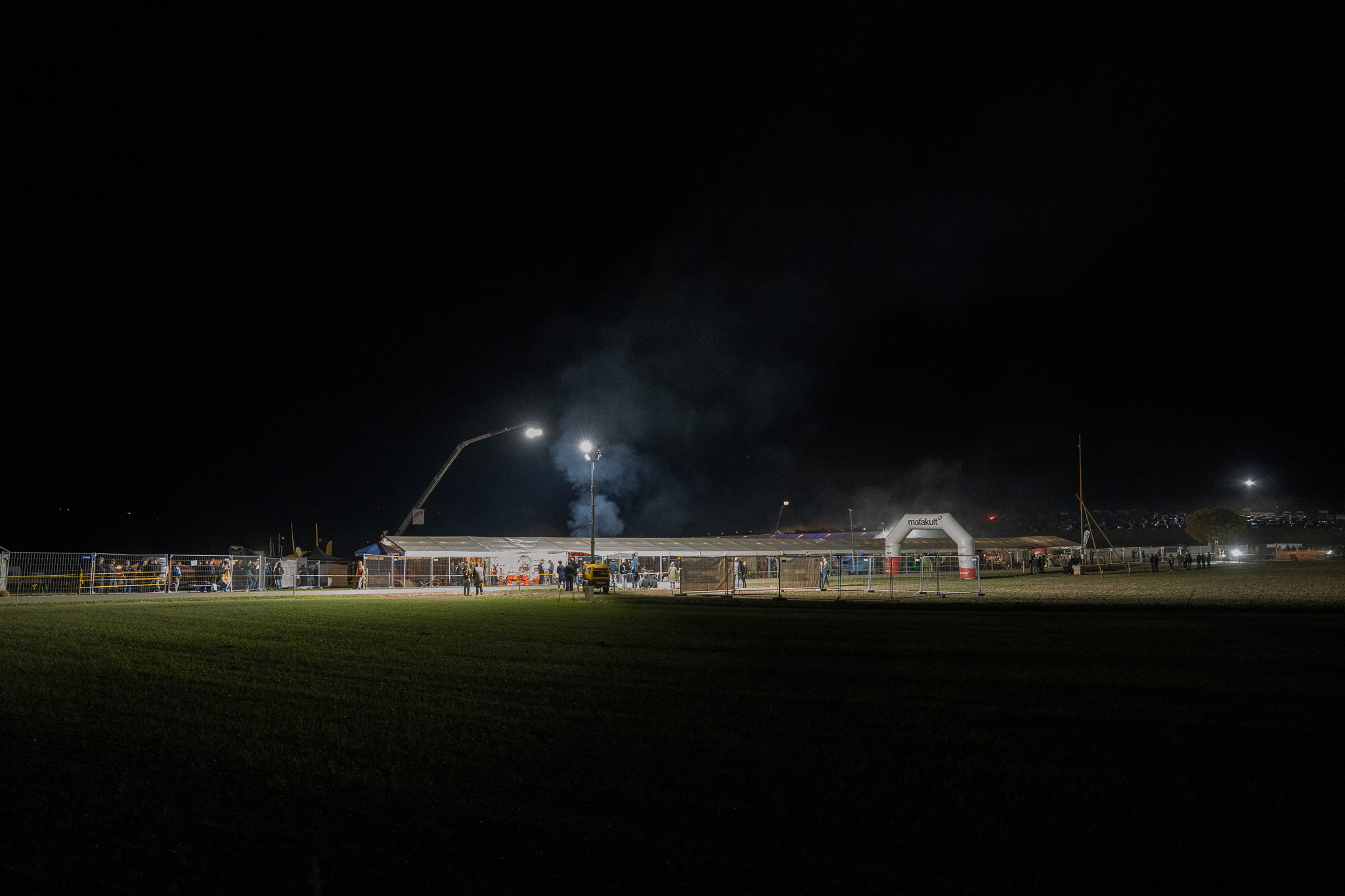 Nachts: Das hell erleuchtete Festzelt am Rennplatz mit Bauzaun, Flutlicht an einem Kran, aufsteigendem Rauch und einem aufblasbaren Torbogen, davor eine dunkle Wiese