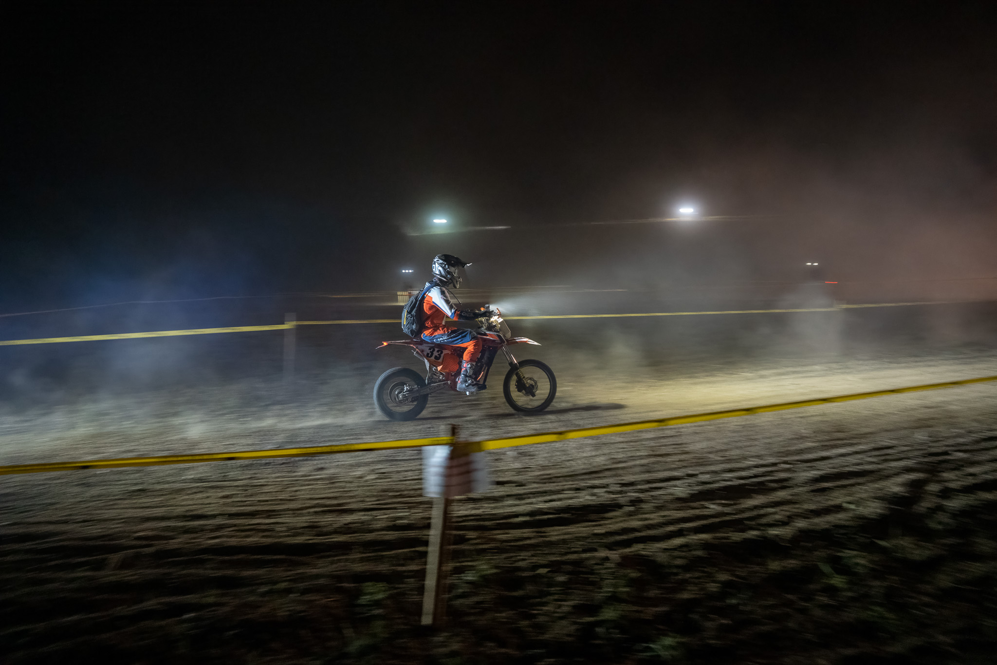 Nachts: Fahrer mit Startnummer 33 und Rucksack rast durch Staubschwaden, im Hintergrund die Flutlichtmasten der Rennstrecke, vorn das gelbe Absperrband