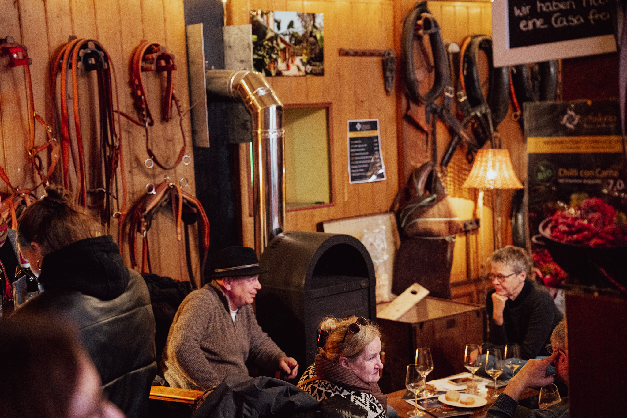 Gäste mit Weingläsern an Holztischen in der Sattelkammer, an der Wand hängen Trensen, Zaumzeug und Pferdegeschirre