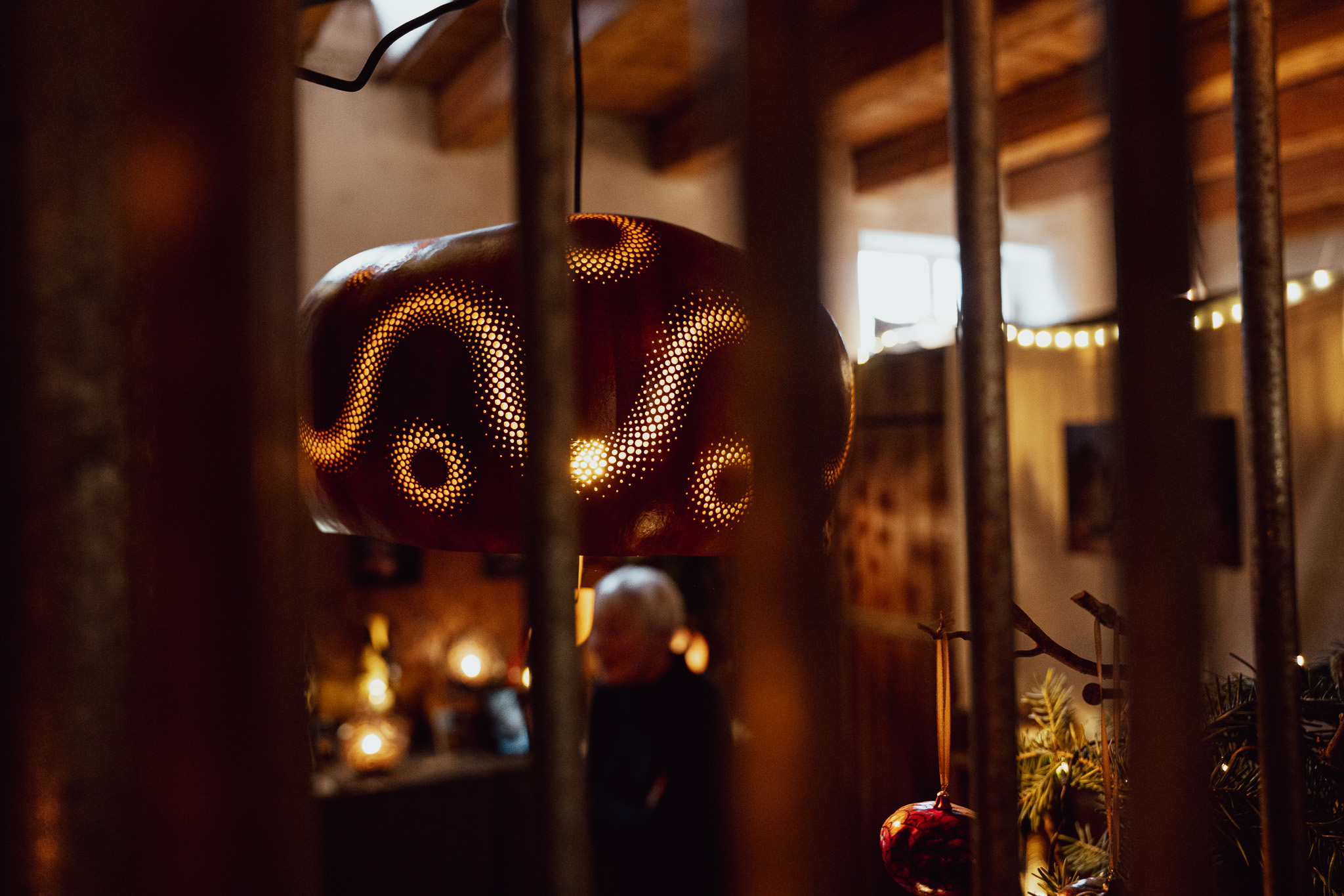 Durchbrochene Keramiklampe mit warmem Licht hinter einem Stallgitter: die gelochten Muster werfen Punkte aus Licht, dahinter Kerzen und Christbaumkugeln