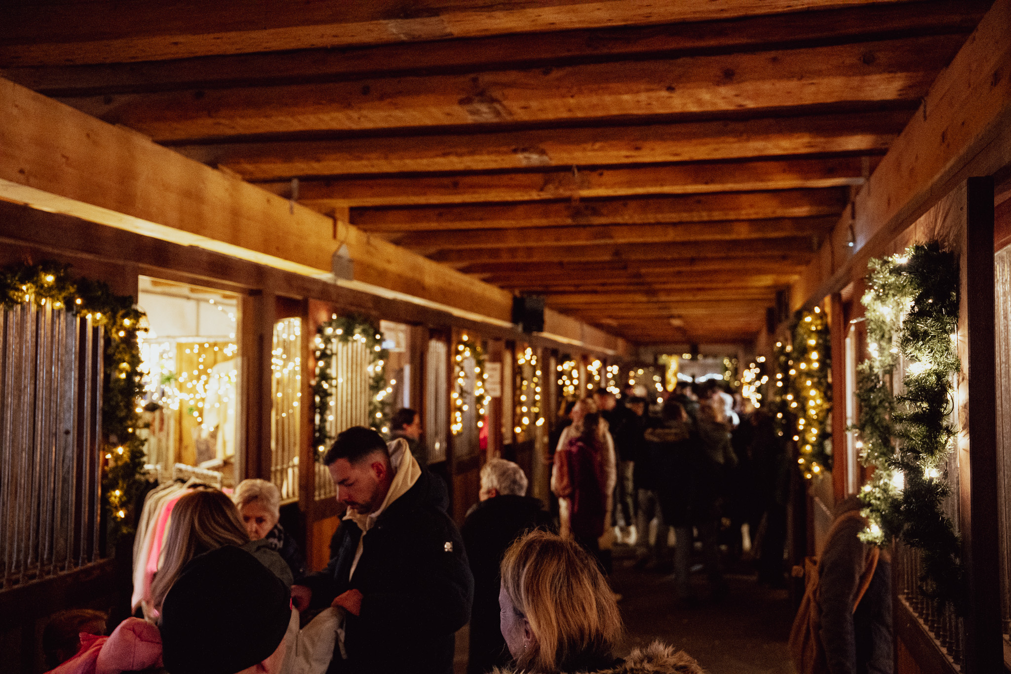 Stallgang mit Holzbalkendecke: an den Pferdeboxen hängen Lichterketten und Tannengirlanden, Besucher stöbern an den Ständen in den Boxen