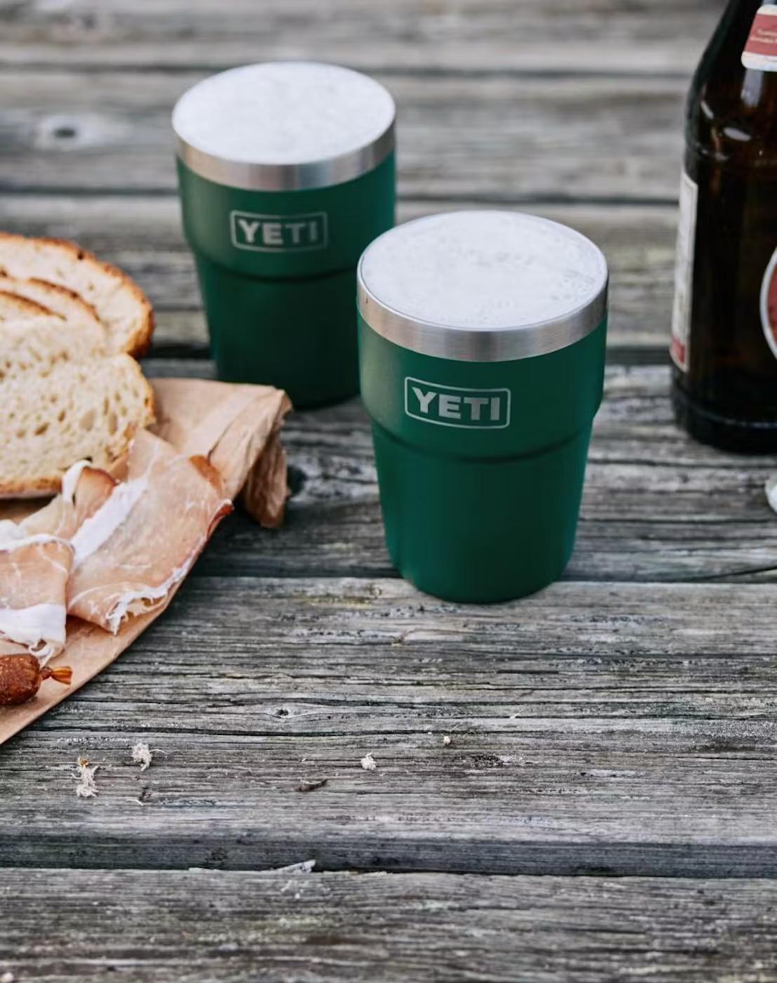 Deux mugs YETI vert foncé avec de la mousse de bière sur une table en bois patinée, à côté du pain tranché, du jambon sur papier et une bouteille de bière
