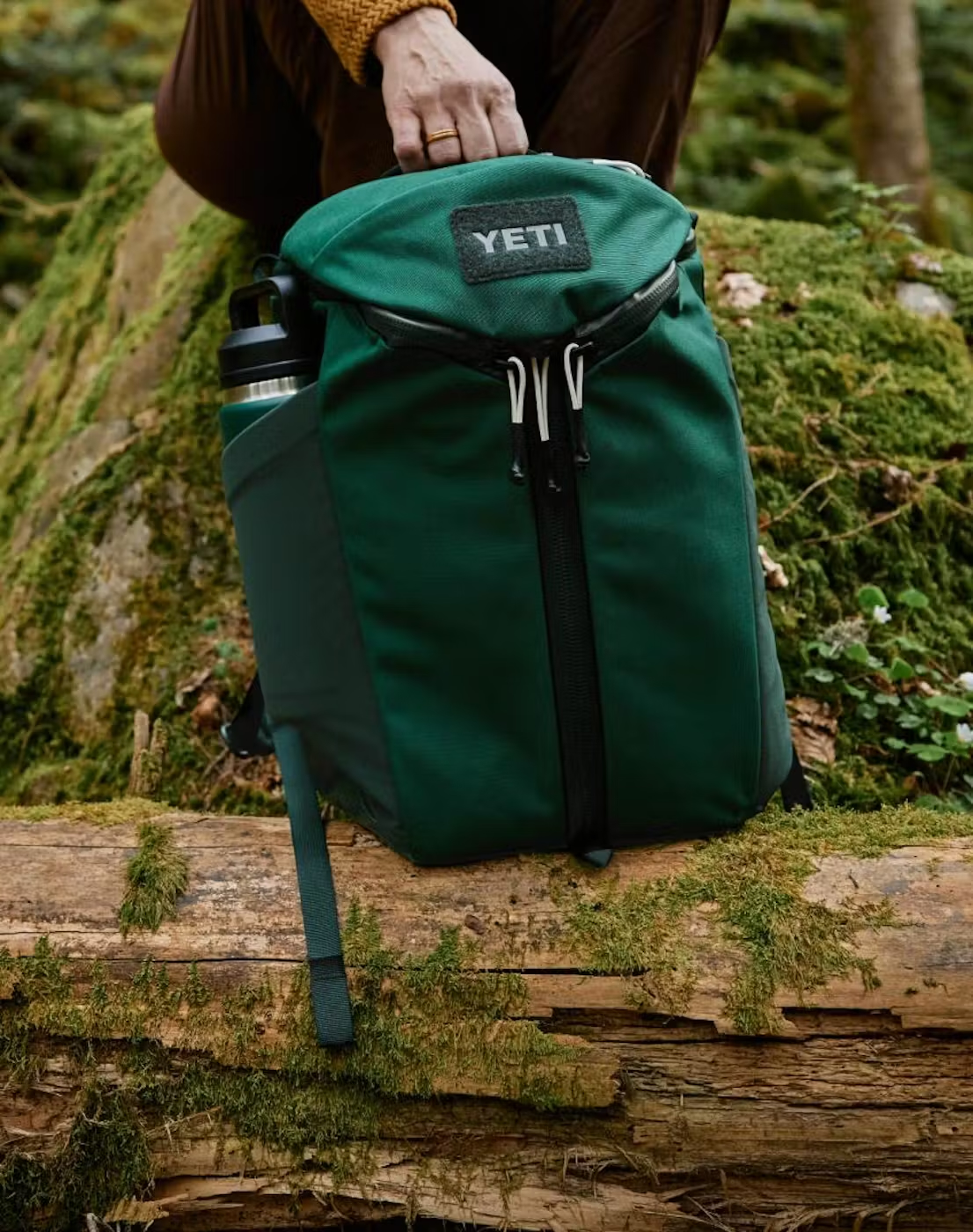Une main soulève d'un tronc moussu un sac à dos YETI vert foncé avec une gourde dans la poche latérale